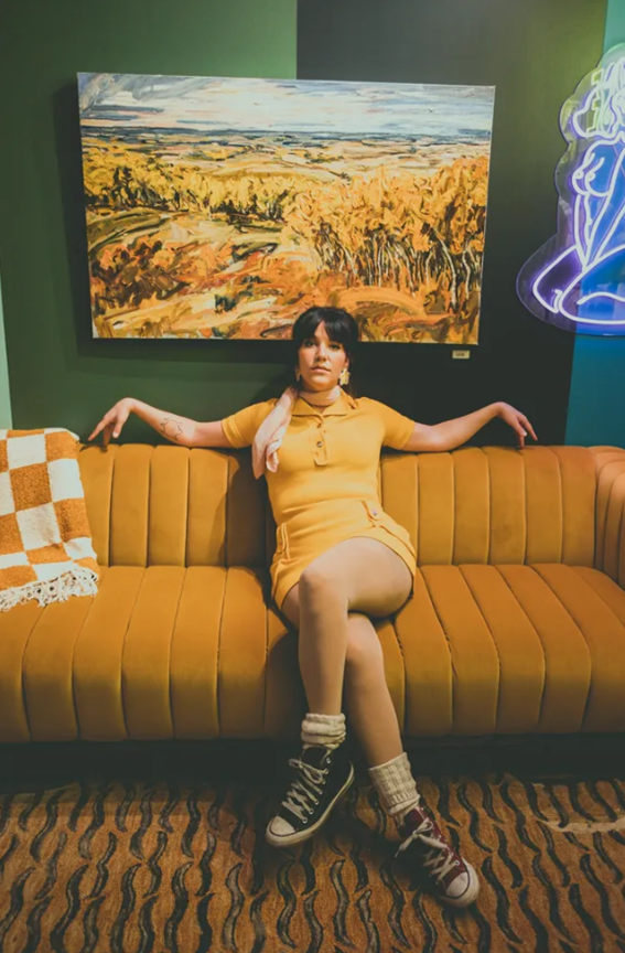Dakota Ray Hebert - Saskatchewan artist and Indigenous woman in a retro-styled photo sitting in a yellow couch with checkered white and a yellow blanket hanging on the back. The woman in wearing a short yellow dress with, sheer/whiet pantyhose, white socks and converse sneakers, black on her left foot and red on her right foot. Her elbows are resting on the back of the couch. Photo on SK Arts website courtesy of the artist.
