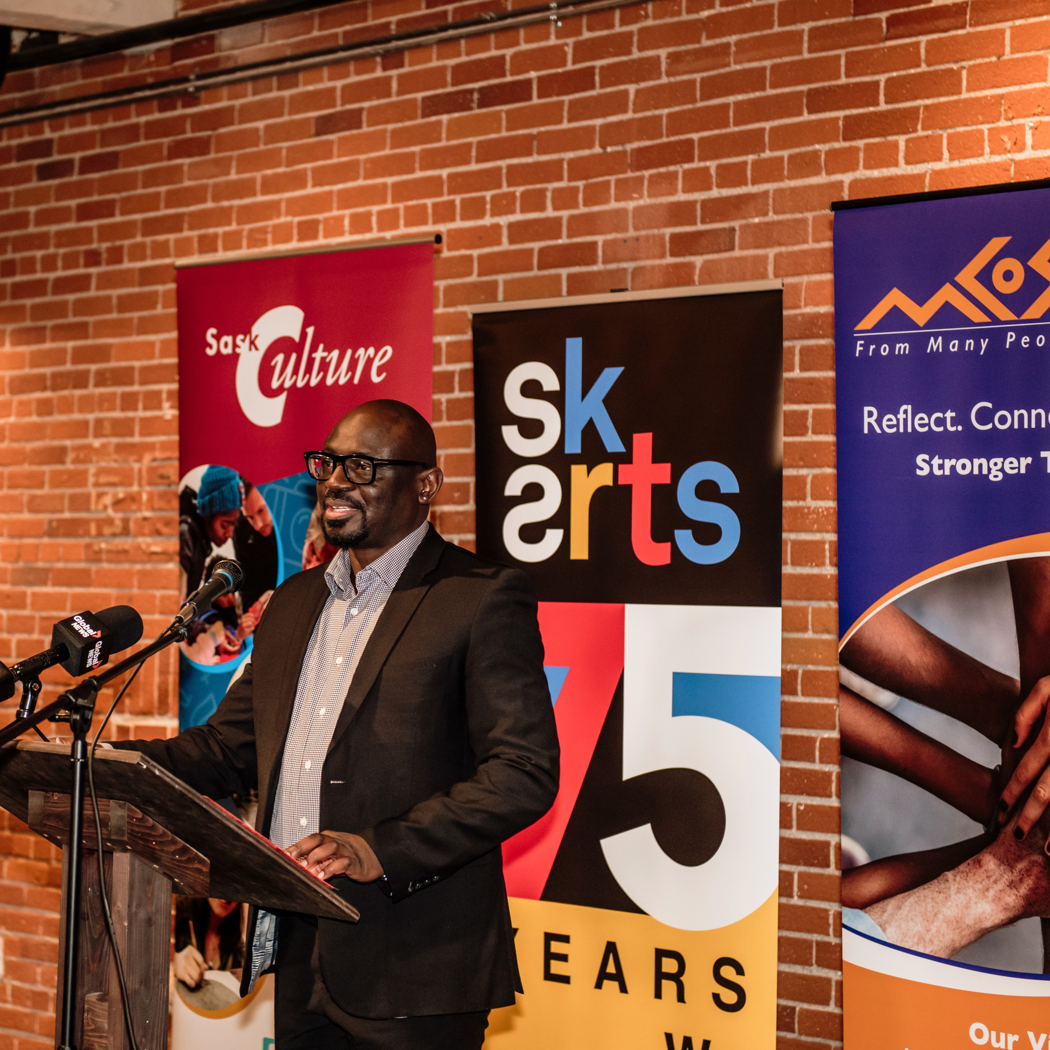 Black man at a podium with microphone making a speach. There is brick wall and banners behind him