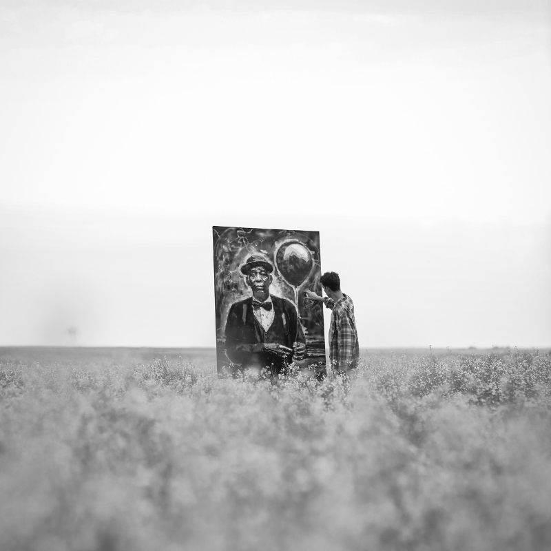 Andrew Robertson - Canadian Artist, Shaunavon Saskatchewan - Black and white photo of a man in the distance in a field in the distance working on a a large black and white painting of a man holding a balloon.