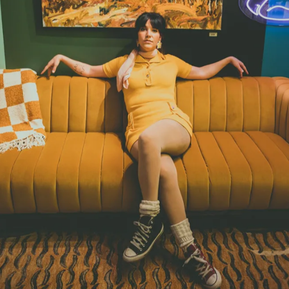 Dakota Ray Hebert - Saskatchewan artist and Indigenous woman in a retro-styled photo sitting in a yellow couch with checkered white and yellow blanket hanging on the back. The woman in wearing a short yellow dress with, sheer/whiet pantyhose, white socks and converse sneakers, black on her left foot and red on her right foot. Her elbows are resting on the back of the couch. Photo on SK Arts website courtesy of the artist.