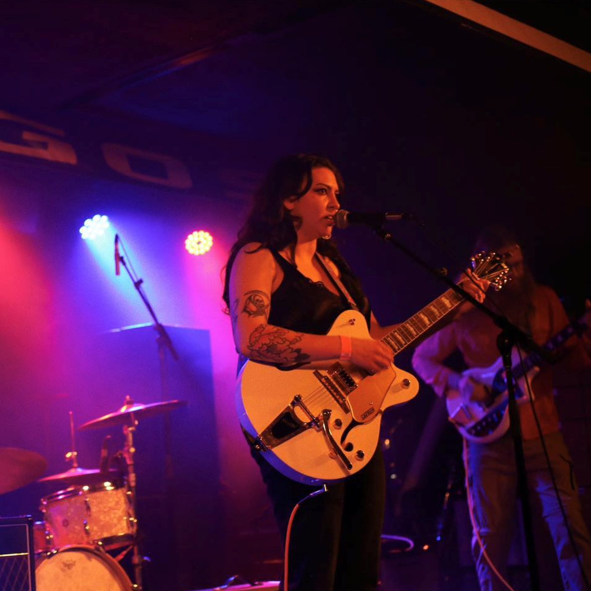 Aurora Wolfe - 75th anniversary nominee, picture shows a woman on stage with long dark hair wearing a black tank top and playing a guitar and singing. One arm has a half sleeve tattoo.