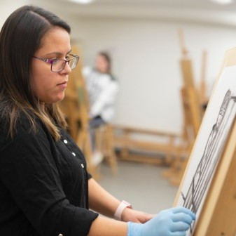 Fine Arts Department - Young woman with brown hair wearing blue gloves drawing on a canvas with chalkcoal