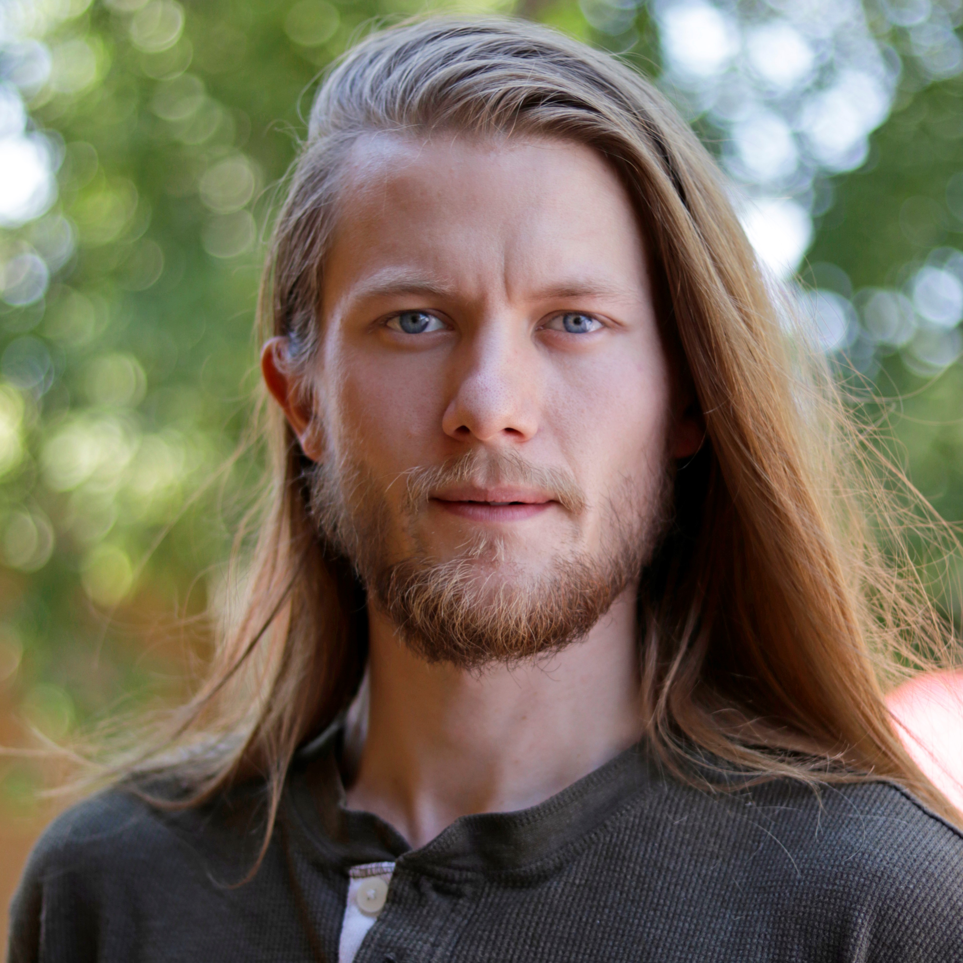 Zac Oliver - Portrait young man with long blonde hair and blue eyes wearing brown shirt.