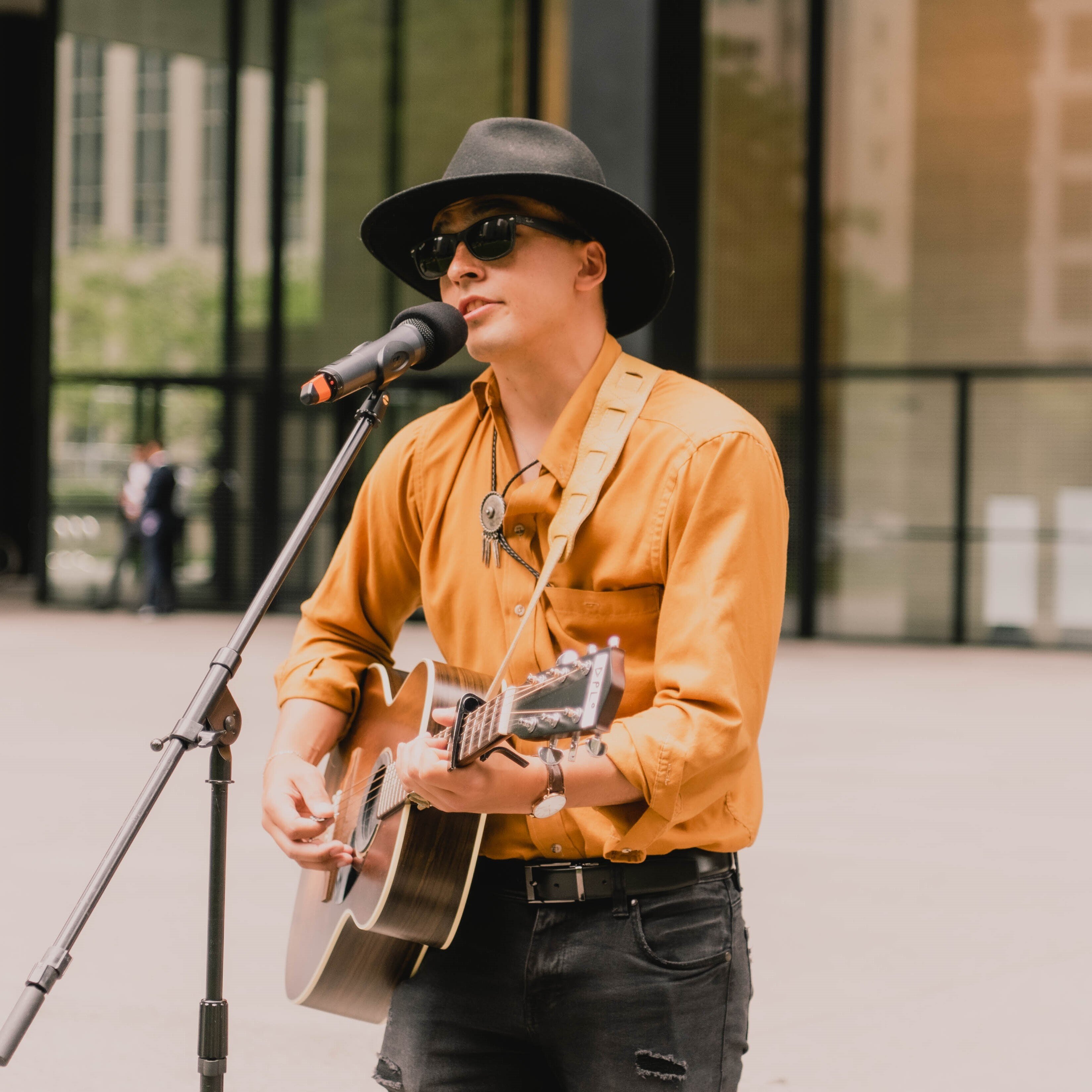 Aren Okemaysim – Indigenous man on stage playing a guitar and singing. He is wearing a hat, orange shirt and sunglasses.