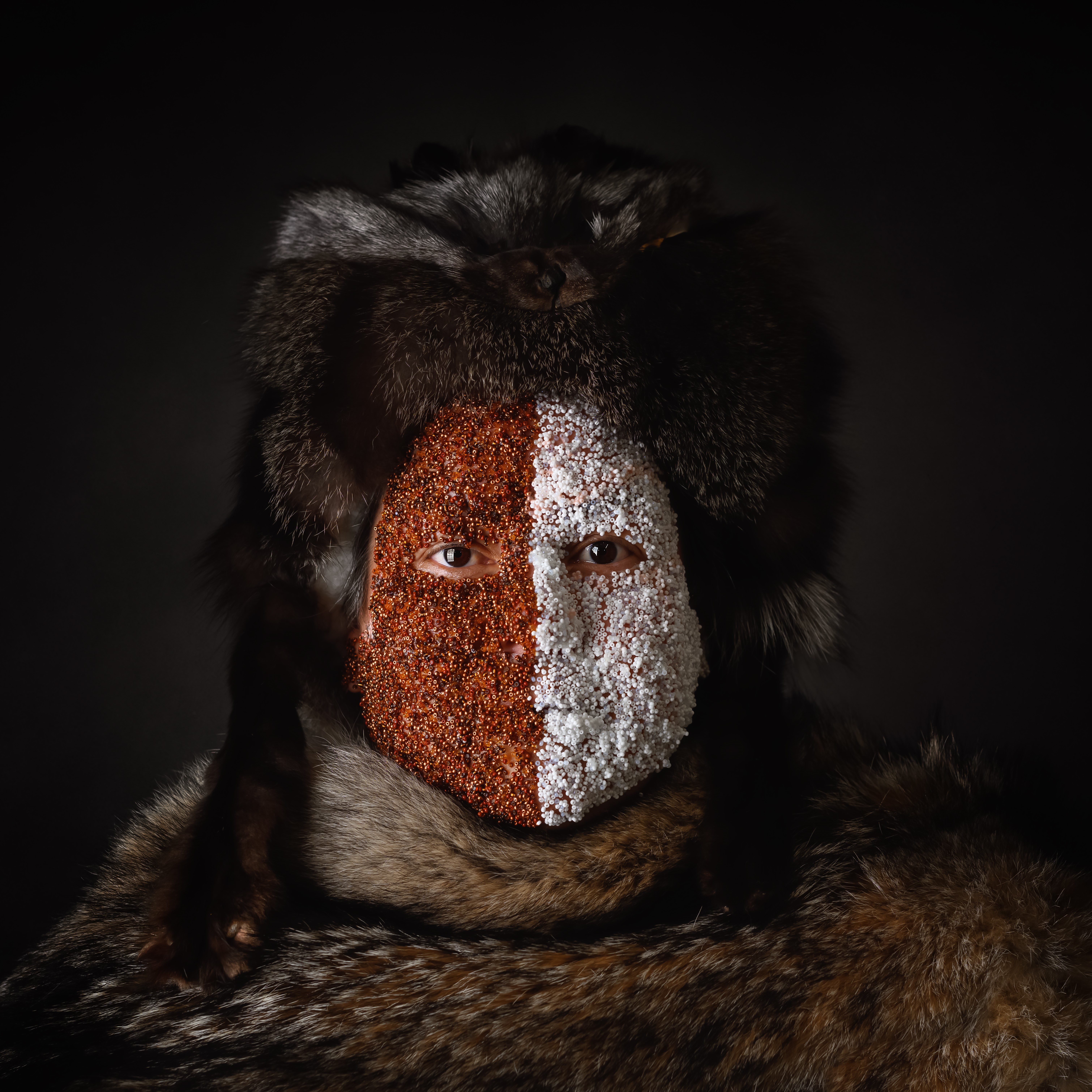 Marcy Friesen - Half Breed - Photo of Indigenous woman wearing fur hat and cloak and white beads on half  of her face and brown beads on the other half.