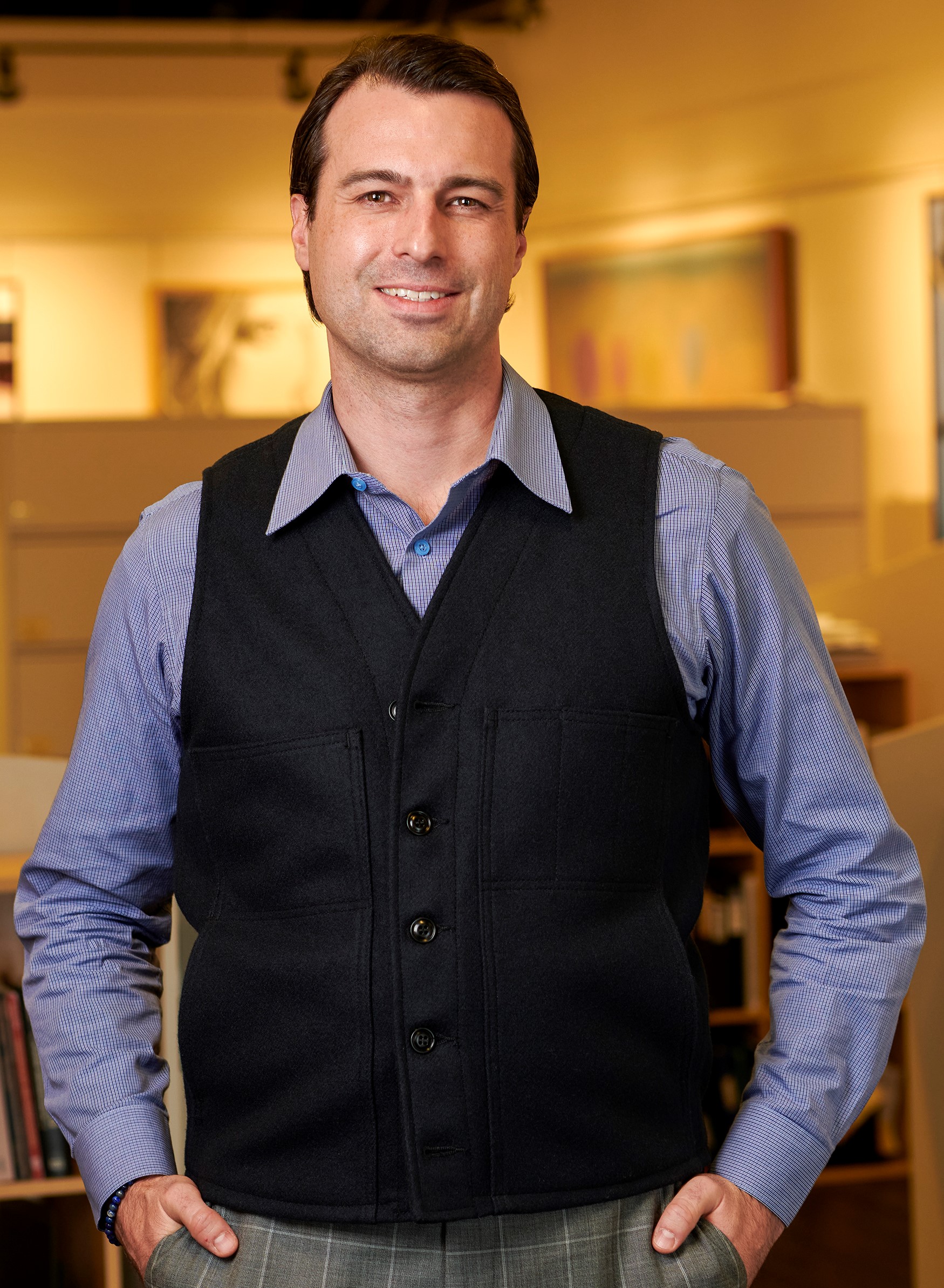 Keith Busch - Tall, white male with brown hair wearing a blue shirt, black vest and grey pants. He smiling,  is  standing in an office, with hand in his pockets. The picture has a blurred background.