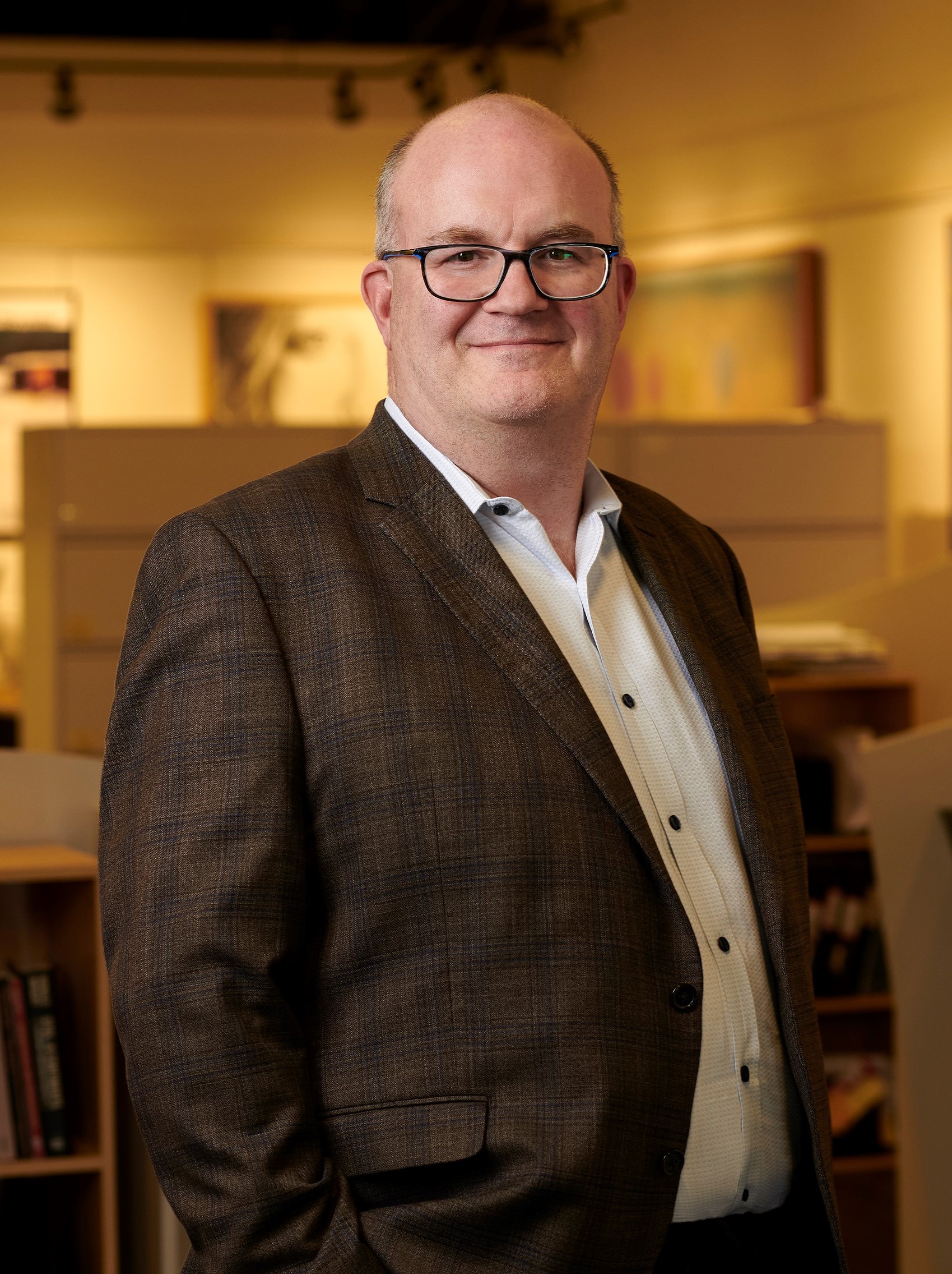 Older, white male with black rimmed glasses wearing a cream shirt and brown jacket. He is  standing in an office with a blurred background smiling.