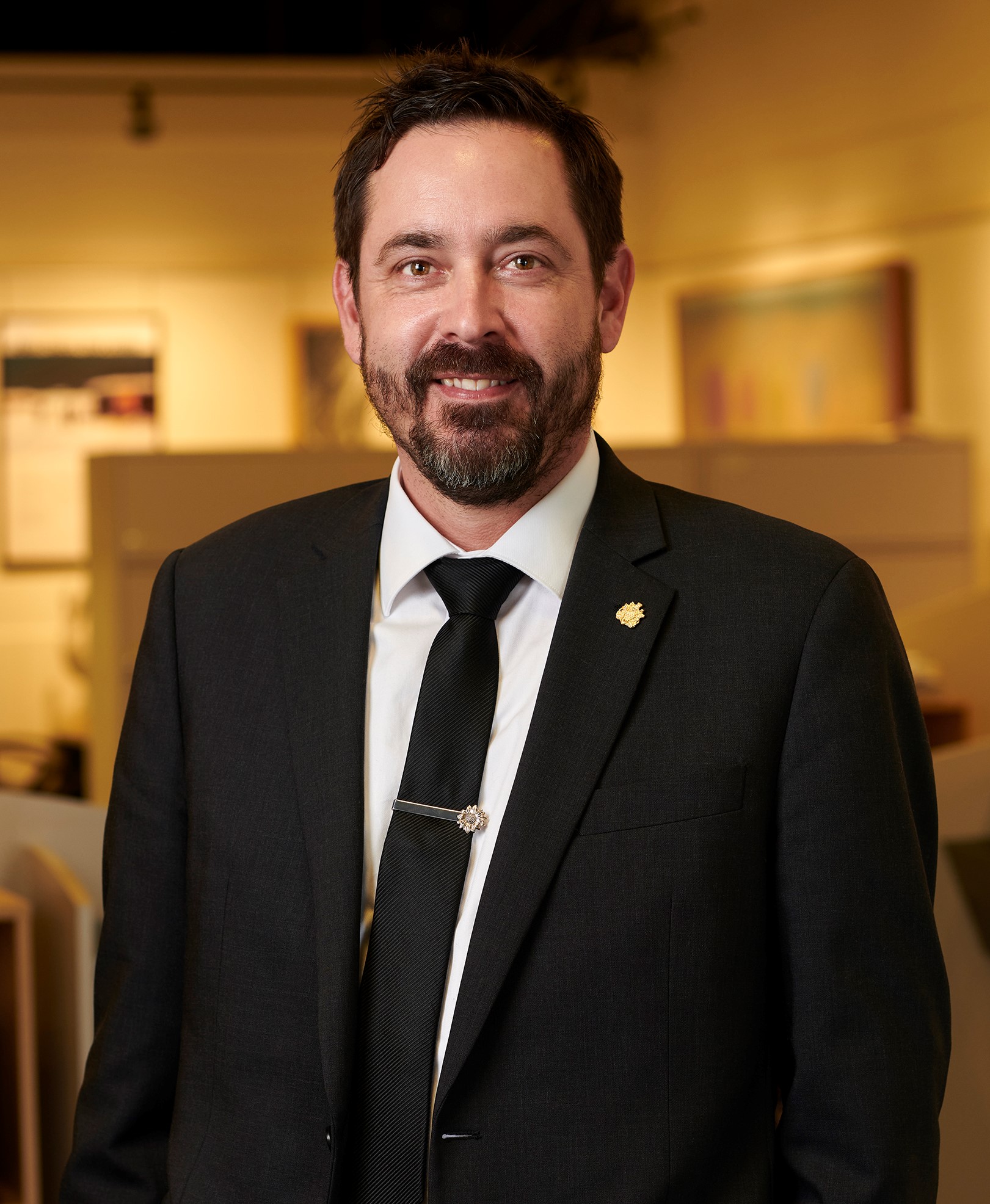 Shawn Blackman - Tall, white male with brown hair and a beard  wearing a black suit. He is  standing in an office with a blurred background smiling.