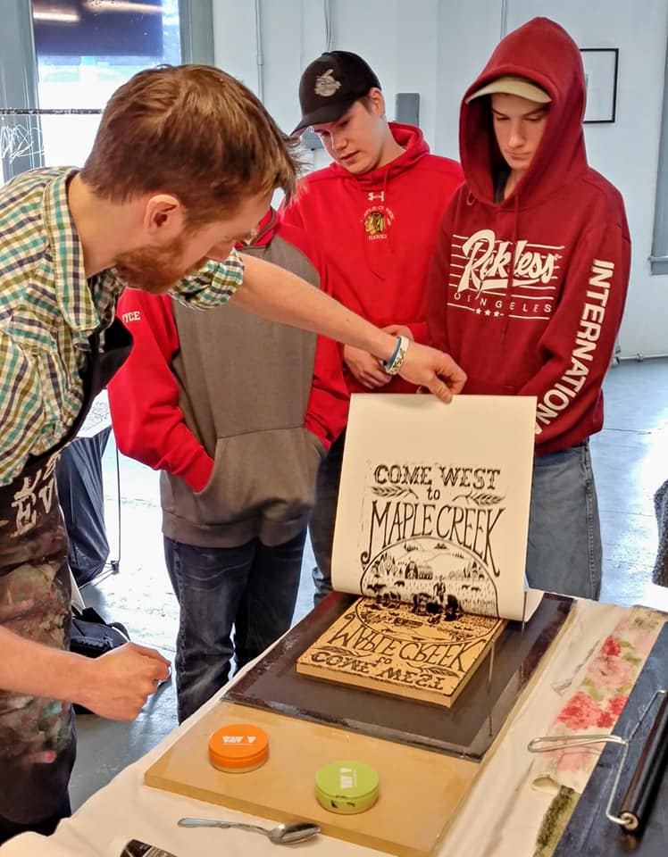 SKArts - rtist Geoff Phillips demonstrates "pulling a print" from a block as a hands-on illustration of the printing of posters at the turn of the last century. Geoff hand carved this print block to represent the style of settlement promotional posters that were common during the Settlement Boom era.