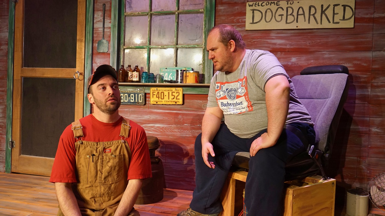 Two men sit on a porch with a sign reading, "Welcome to Dogbarked" hung on the exterior of the house behind them.