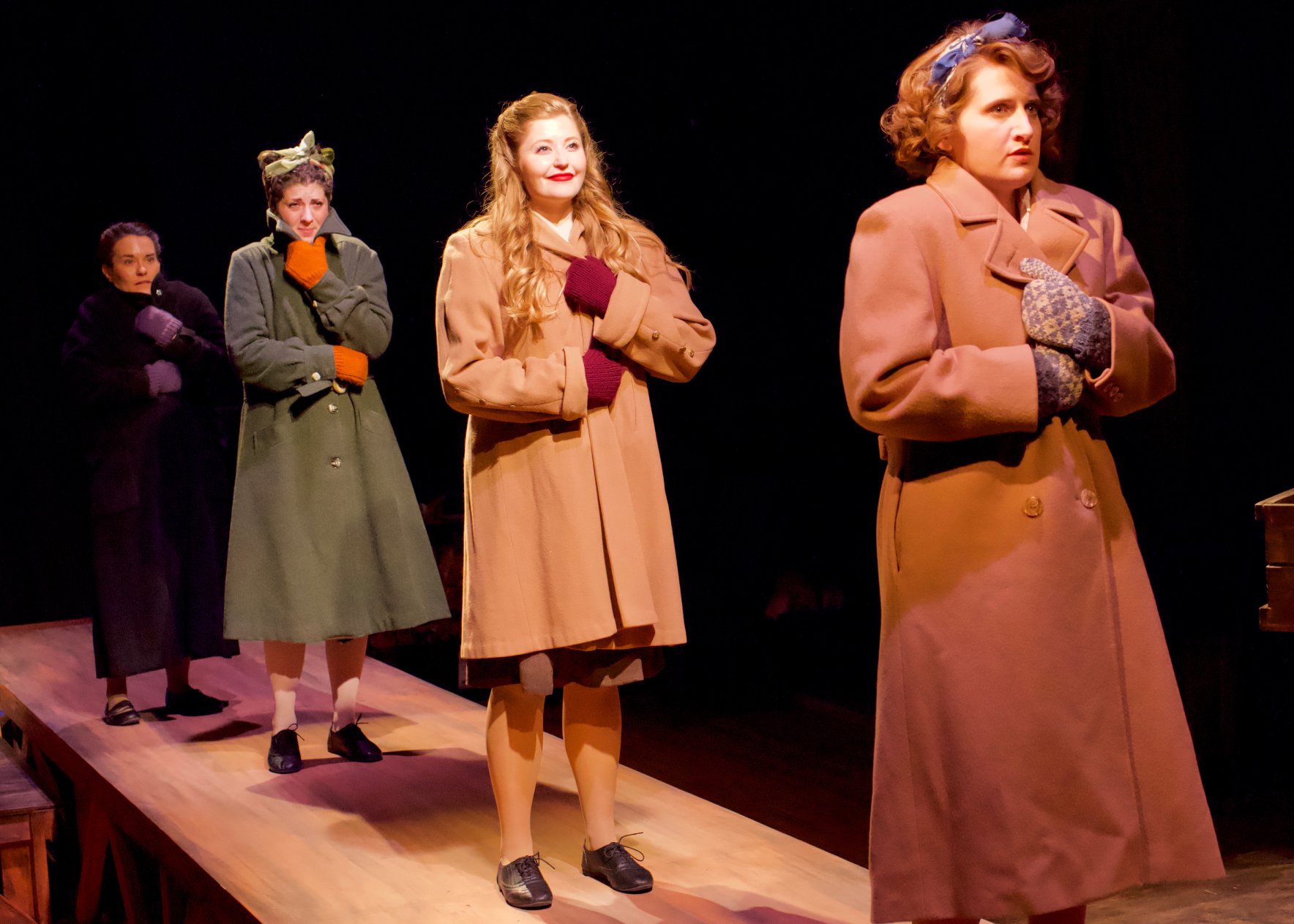 Four women wearing coats stand in a row on a raised platform.