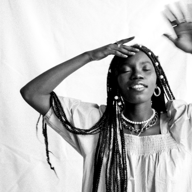Black and white photo, Peace Akintade, black young woman in a dress and braids touching her forehead and holding up and open palm in candid pose