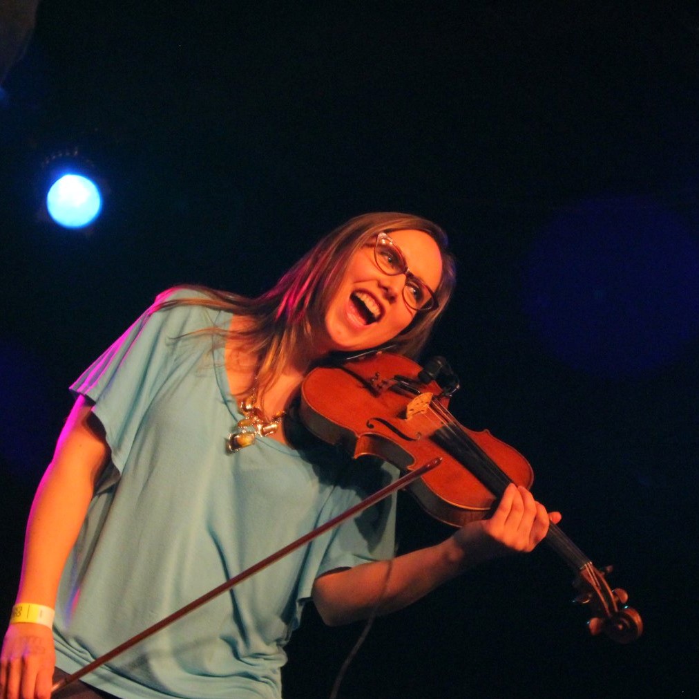 Carmelle Pretzlaw, Adminstrative Coordinator (Programs) - Woman on stage with a violin playing. She is wearing glasses, smiling and wearing a green blouse.