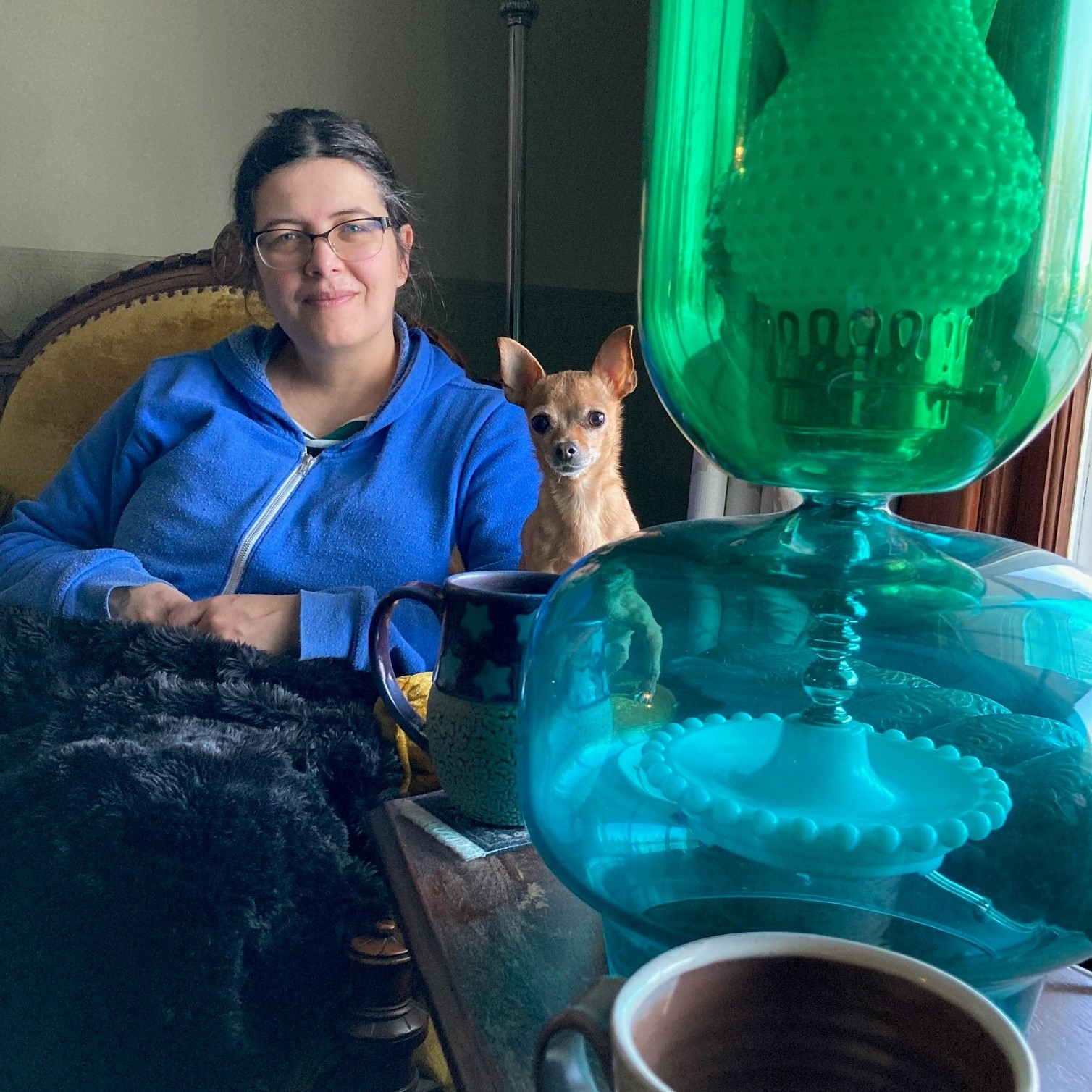 Julia Krueger, SK Arts Registrar (Permanent Collection) - Woman sitting in a chair wearing a blue hoodie and is covered by a dark coloured blanket. In the foreground is a green glass on a table.