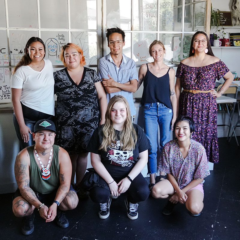 Chokecherry Studios, Saskatoon arts organization. Group of eight people in an office space. Description is from left to right at the back - Young indigenous woman with long hair, wearing white t-shirt and jeans smiling broadly with one hand by her side and the other hugging a young woman to her left. Short indigenous young woman with short orange hair wearing a dress. Young indigenous man wearing glasses and a blue button down shirt and his arms folded across his chest with slight smile. White, blonde young woman wearing a black tank top and jeans. Young indigenous woman with long brown hair and wearing glasses smiling. She is wearing a long, off the shoulder floral dress. Description and then left to right from front - Young indigenous man with tattoos stoops smiling. He is wearing a baseball cap, a dark green tank top and blue jeans shorts. Blonde woman stooped smiling with long blonde hair, black t-shirt with graphics, jeans and sneakers. She has red makeup around her eyes and is smiling. Young woman in floral top stooping. She is of Asian descent, with short black hair.
