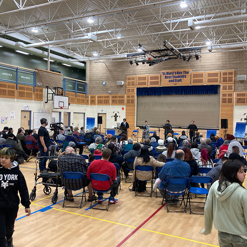 Image of gathering in a basketball court. There are chairs facing a stage with four speakers.
