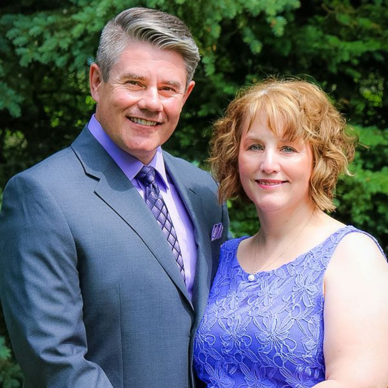 Kevin and Carroll Dyck - Older Man and woman standing closely together. The man has grey hair, is where a grey suit with lavender shirt and purple tie. The woman in wearing a purple dress.