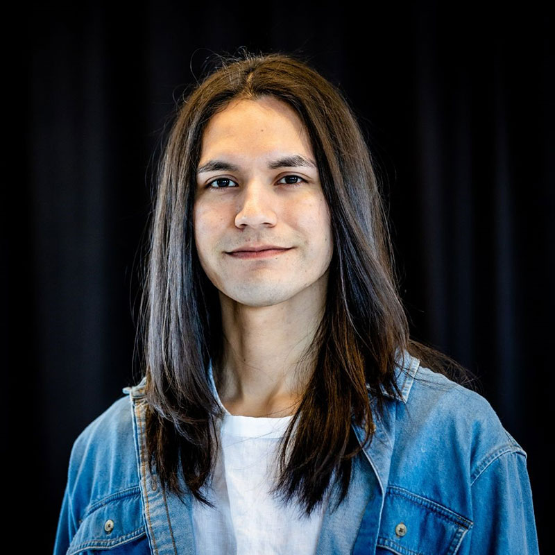 Marcus Merasty - Headshot of young man with long black hair smiling. He is wearing a jean jacket and white t-shirt