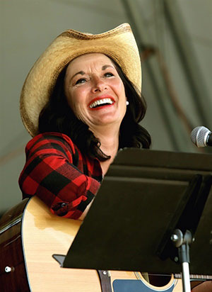 Jeanette Wicinski Dunn at Country at the Creek for SK Arts Arts of Celebration 2025. Woman on stage playing guitar and smiling on stage wearing large country has and red flannel shirt.
