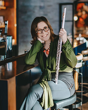 Marie Véronique Bourque for SK Arts Arts of Celebration 2025. Woman with a musical instrument sitting at a bar and smiling.