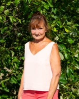 Photo of woman with brown hair and bangs standing in fron tof green trees, wearing a white sleeveless blouse and smiling. - Lynda Monahan no credit